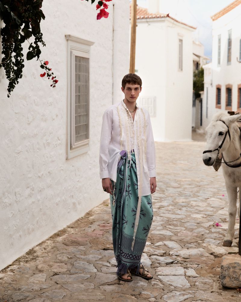 Young man in a white shirt and patterned skirt posing on a cobblestone street next to a white donkey in a sunny, outdoor setting.