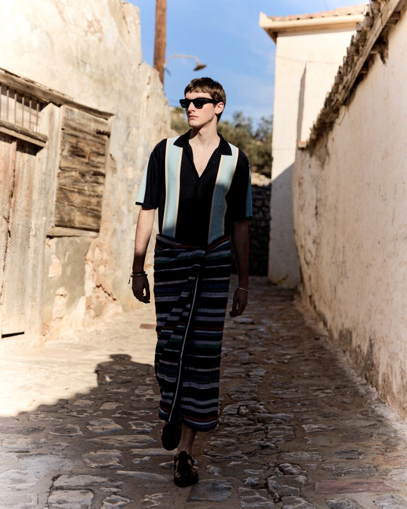 A stylish man wearing sunglasses walks down a cobblestone street flanked by old buildings on a sunny day, showcasing a unique fashion style.