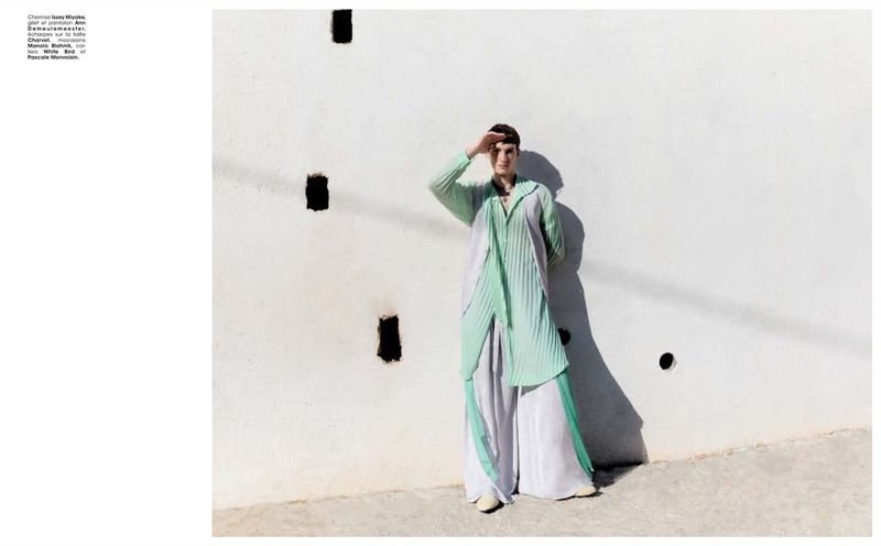 Adult female model poses in mint green pleated outfit in front of a white stucco wall with square holes on a sunny day outdoors.