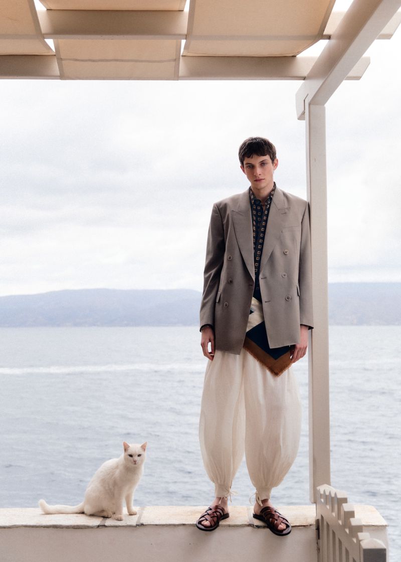 Man in a suit and white balloon pants poses outside by the ocean with a white cat, standing on a wall under a trellis.