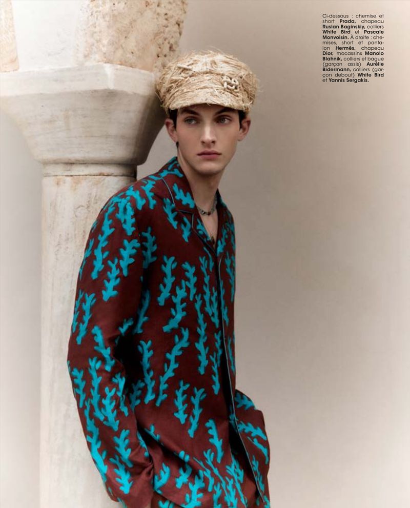 Editorial fashion shot of a young adult man in a patterned shirt and straw cap leaning against a column with neutral background.