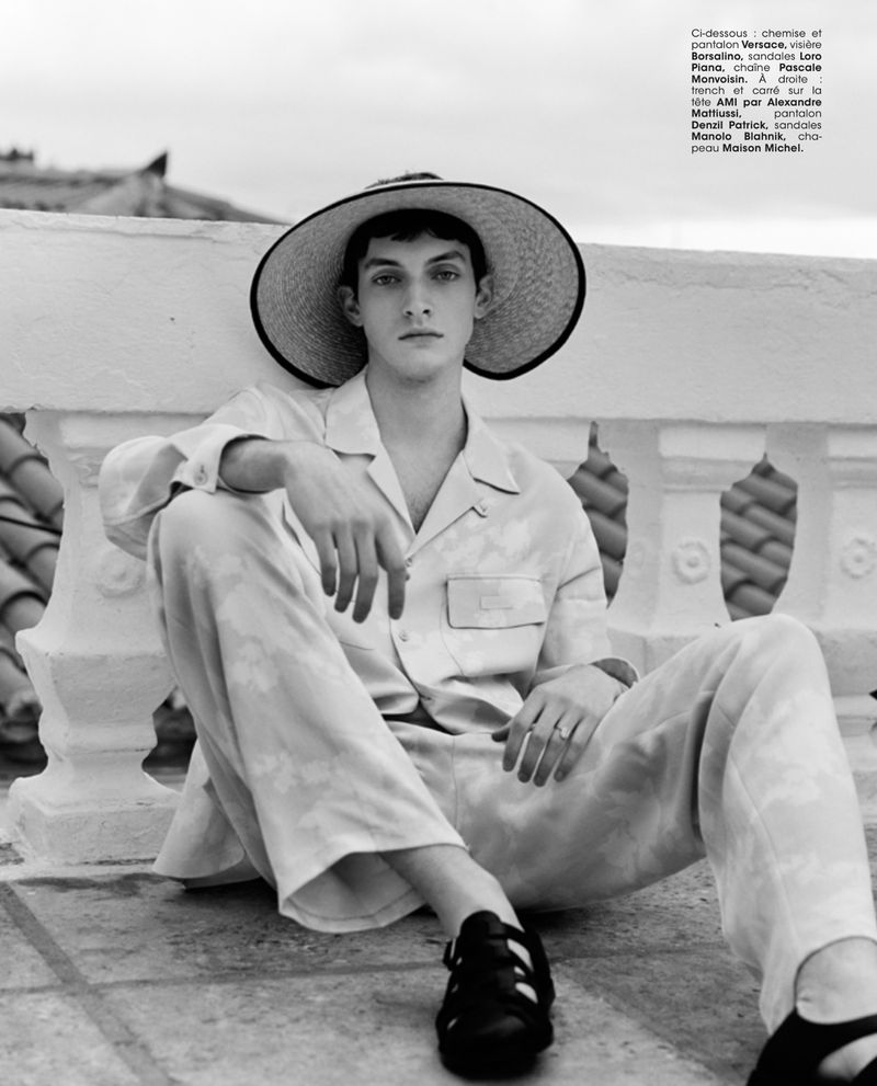 Black-and-white fashion shot of a young adult male wearing a straw hat, floral-patterned pajama set, and black sandals, sitting outdoors.