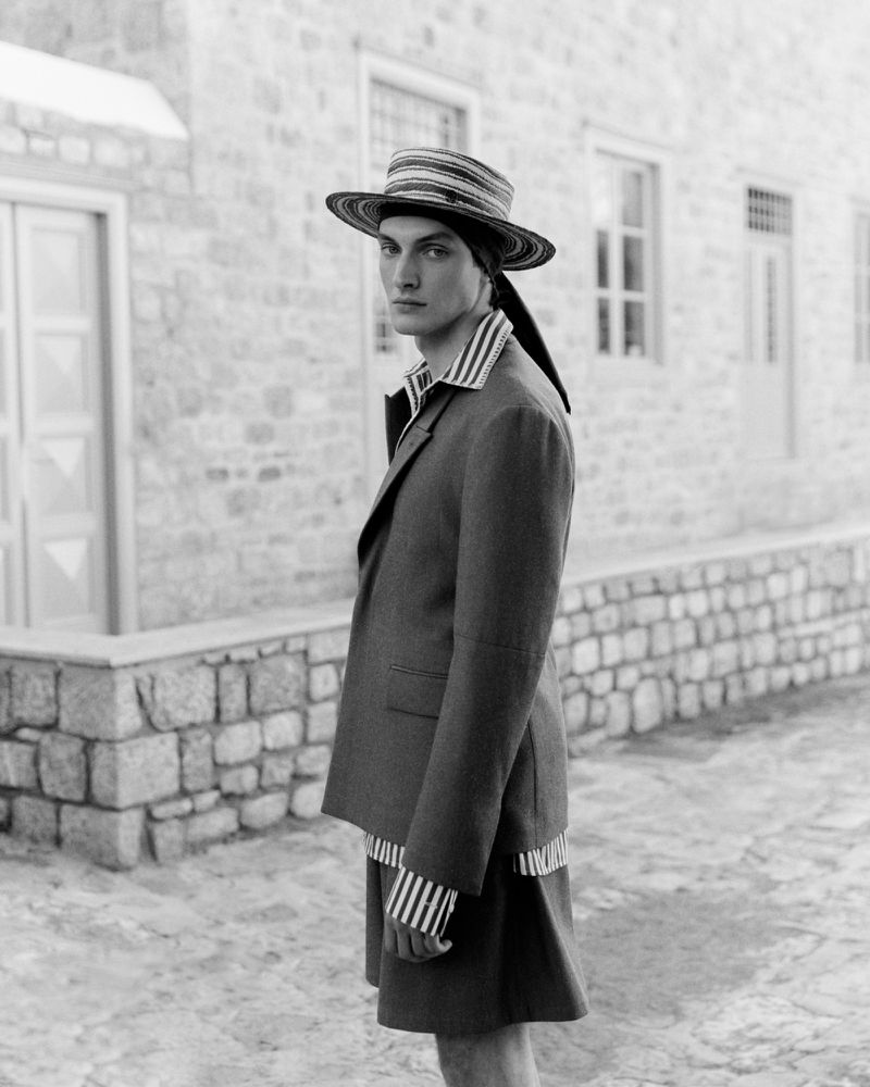 Monochrome fashion shot of a male model in a blazer, striped shirt, shorts, and hat, posing outdoors against a stone building.