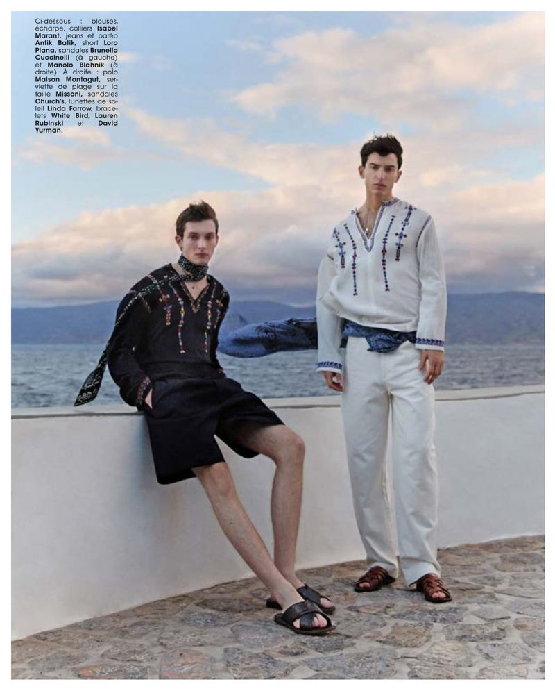 Two male fashion models pose outdoors in embroidered shirts and sandals with a coastal, mountainous background under a cloudy sky.