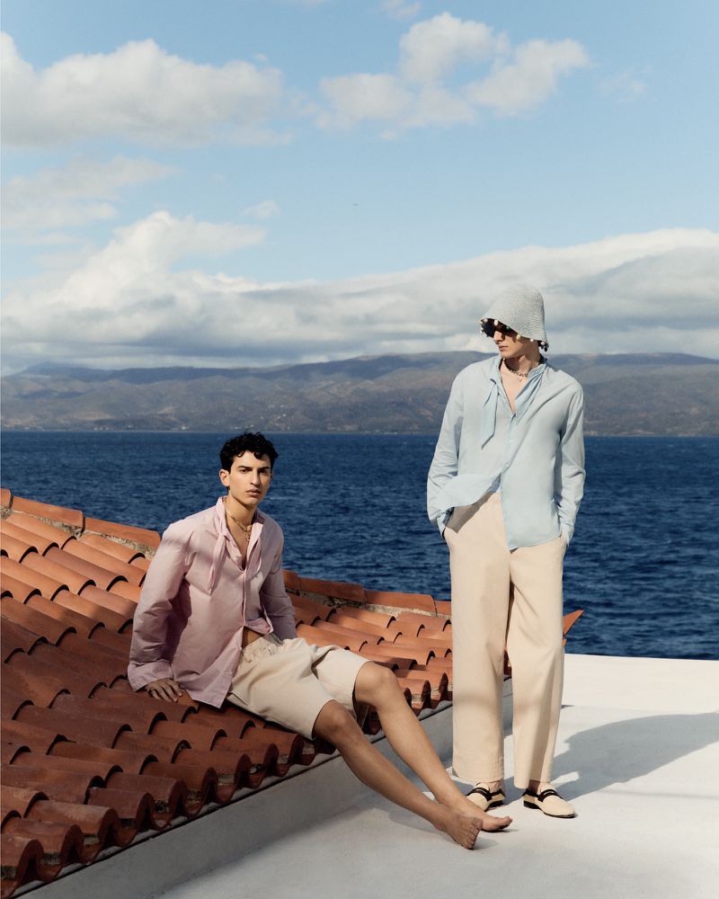 Two adult male fashion models pose on a rooftop overlooking a blue sea and mountainous landscape under a partly cloudy sky on a sunny day.