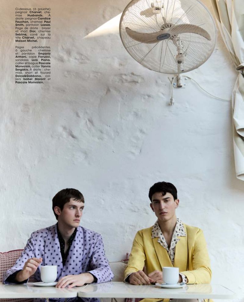 Two young men sit at a table in a bright indoor setting, dressed in patterned shirts and jackets with drinks, beneath a wall fan.