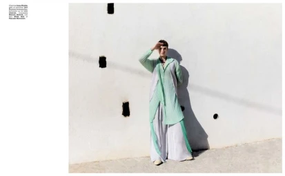 Model wearing a pleated mint green tunic and light grey trousers standing against a textured white wall outdoors.