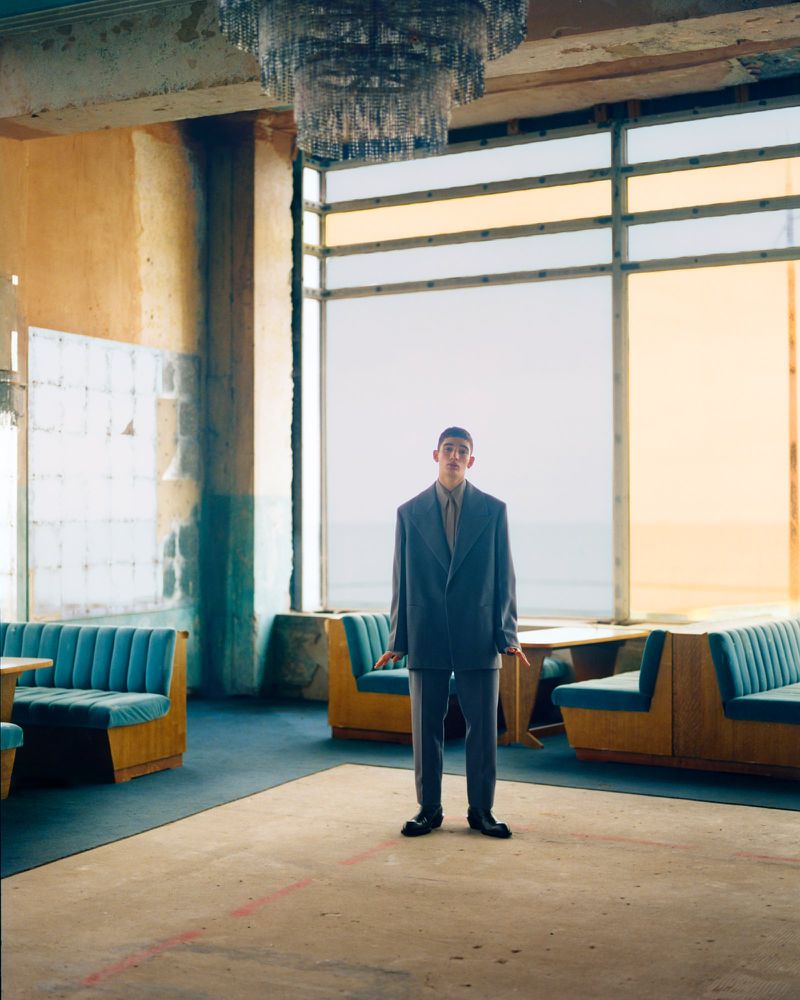 A man in a grey suit stands in an empty hall next to a large window with yellow glass; a chandelier hangs overhead in the abandoned space.