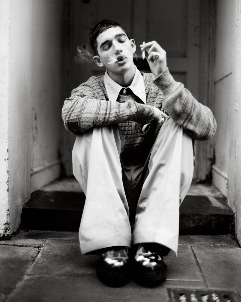 Young man sits with crossed arms, smoking a cigarette in front of a doorway, wearing a sweater, dress pants, and glossy dress shoes.