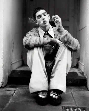 Young man sits with crossed arms, smoking a cigarette in front of a doorway, wearing a sweater, dress pants, and glossy dress shoes.