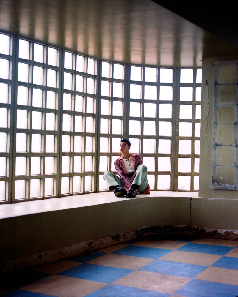 Adult male wearing casual pink jacket sitting cross-legged on a window ledge in an empty room with a blue and beige tile floor.