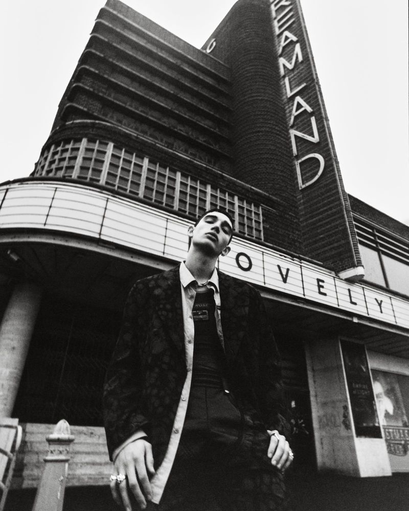 A young adult man in a patterned jacket stands in front of the vintage Dreamland building in a monochrome, low angle fashion shot.