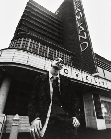 A young adult man in a patterned jacket stands in front of the vintage Dreamland building in a monochrome, low angle fashion shot.