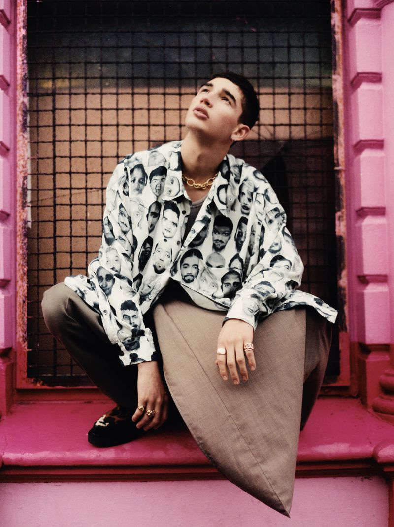 A young man poses in front of a metal gate wearing a button-down shirt with faces printed on it, grey pants, and loafers.