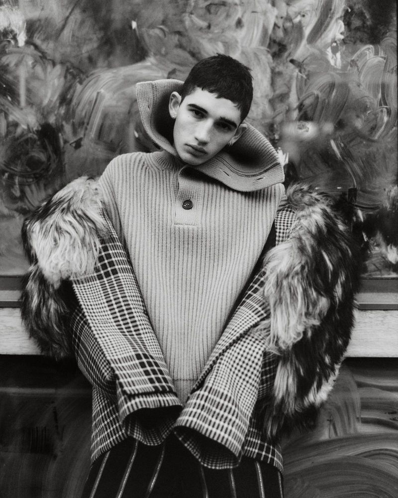 Monochrome portrait of a young male model wearing a high-collared ribbed sweater and a patterned coat with fur trim.