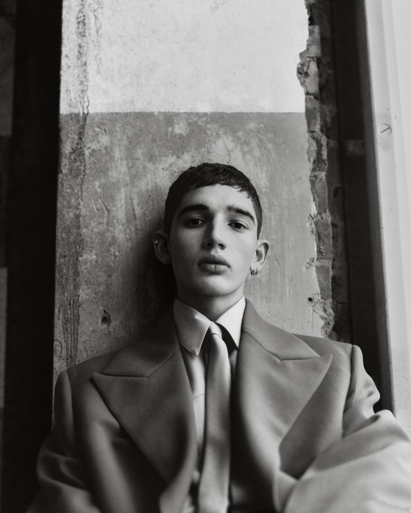 Portrait of a young man in a suit and tie, posing against a textured wall, showcasing a blend of modern fashion and classic style.