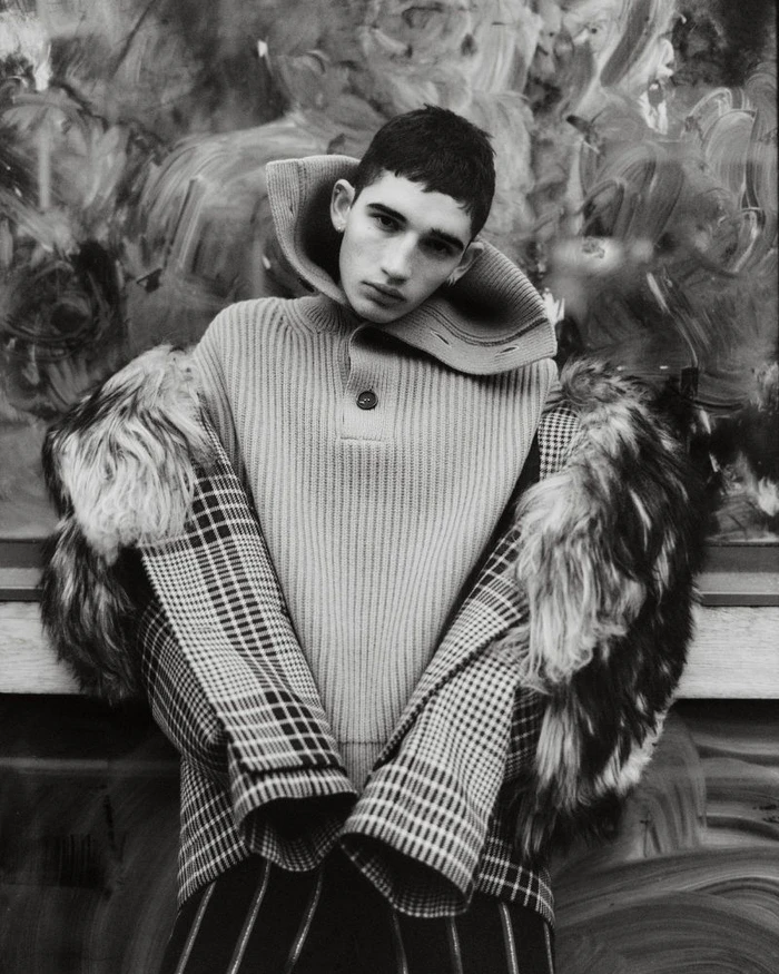 Monochrome portrait of a young male model wearing a high-collared ribbed sweater and a patterned coat with fur trim.