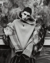 Monochrome portrait of a young male model wearing a high-collared ribbed sweater and a patterned coat with fur trim.