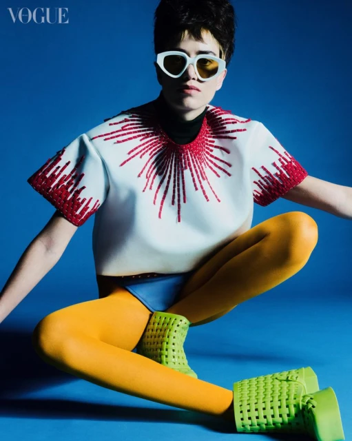 Fashion portrait of a model with short hair, wearing sunglasses and colorful contemporary clothing, in a studio with a blue backdrop.