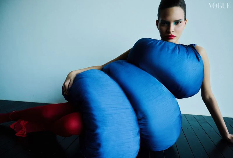 Fashion model poses in a striking blue sculptural dress with red tights against a plain backdrop in a studio fashion editorial.