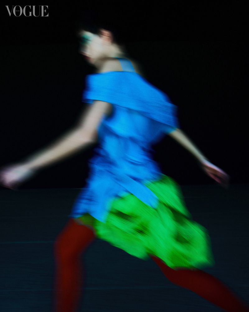 Model in motion wearing a blue top, green skirt, and red tights against a dark background for Vogue Brasil Caleidoscopio project.
