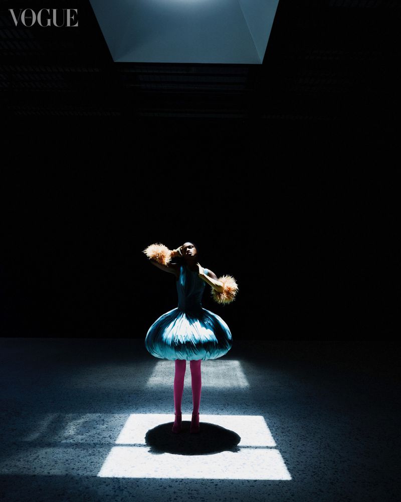 A woman in a blue dress poses under a spotlight in a dark room, wearing furry armbands, with the Vogue logo visible in the top corner.
