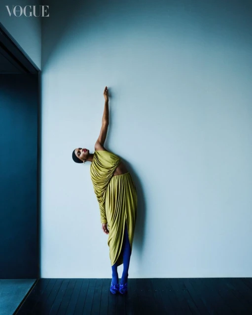 A woman poses in a draped dress and blue shoes in a fashion shot against a blue-grey wall with dark grey floor in a magazine layout.