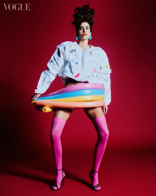 Fashion editorial shot of an adult female model in pink tights and a white jacket holding a colorful inflatable pool toy on a red backdrop.