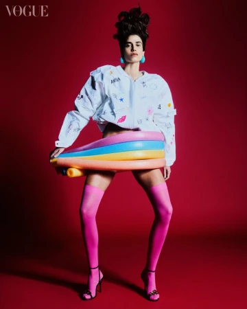 Fashion editorial shot of an adult female model in pink tights and a white jacket holding a colorful inflatable pool toy on a red backdrop.