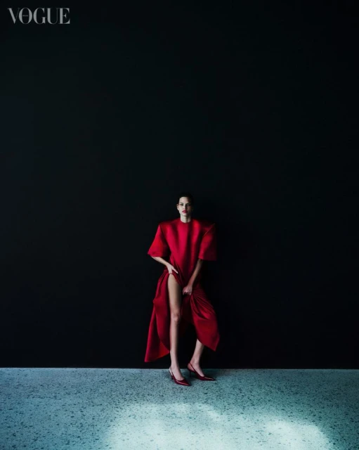 A woman in a red dress posing against a dark background in a fashion editorial. Vogue logo is visible at the top left.