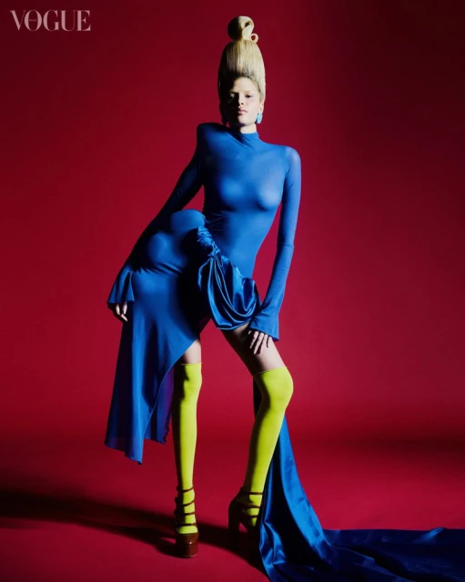 Fashion editorial shot of a model in a blue dress with yellow stockings against a red backdrop, showcasing a high-fashion look.