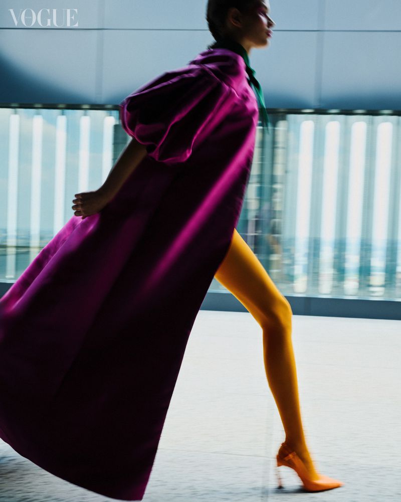 Woman walks in purple coat, yellow pants, and orange heels against an abstract blue background in a fashion editorial image.