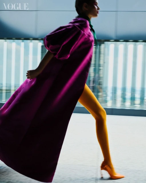 Woman walks in purple coat, yellow pants, and orange heels against an abstract blue background in a fashion editorial image.
