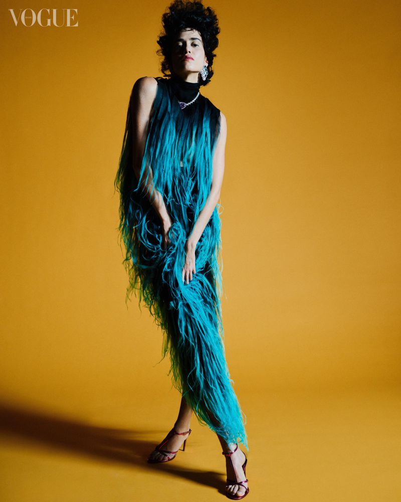 A woman poses in a feathered teal gown in a full shot set against a yellow backdrop for a fashion editorial, showcasing a modern and avant-garde style.