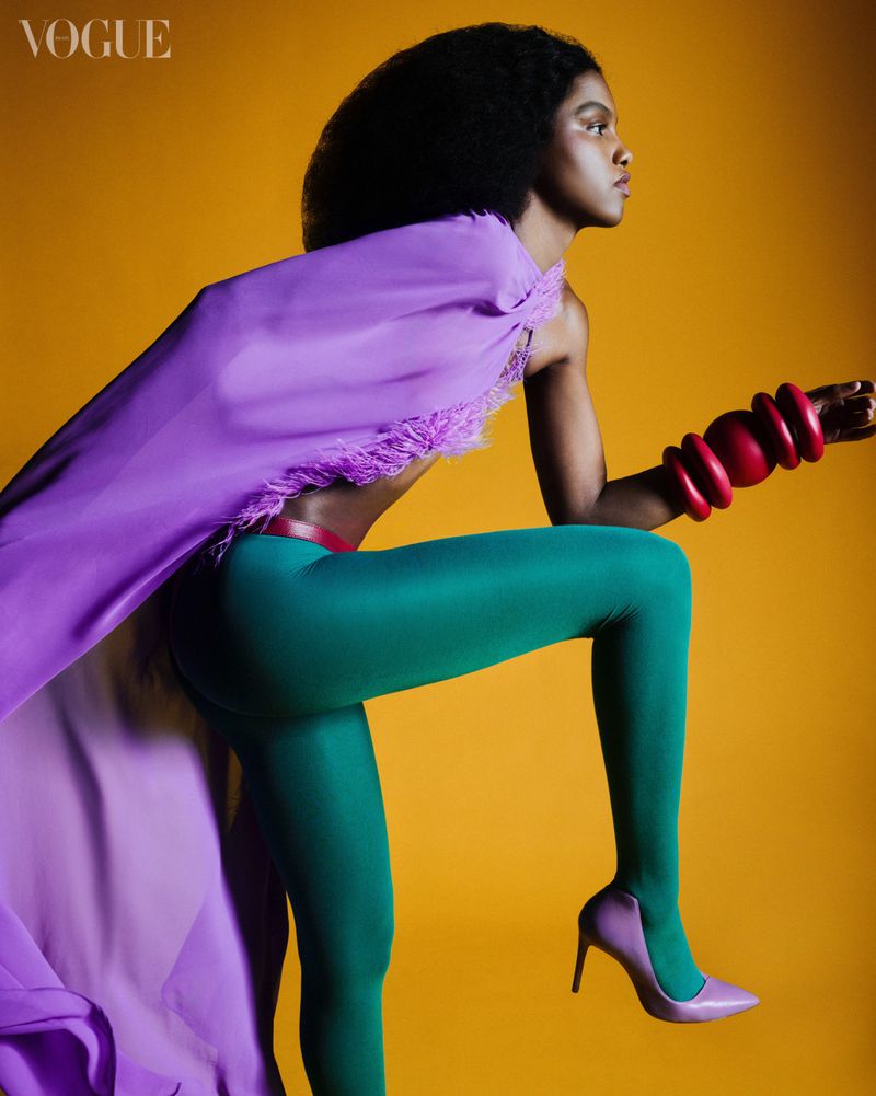 Fashion shot of a Black female model in green leggings, purple heels and top, and red bracelets striking a dynamic pose on a yellow backdrop.