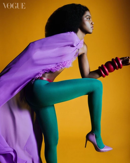Fashion shot of a Black female model in green leggings, purple heels and top, and red bracelets striking a dynamic pose on a yellow backdrop.