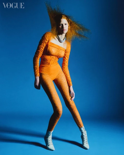 Fashion editorial shot of a redhead female model in an orange lace bodysuit and white high-heeled boots in a blue photo studio.