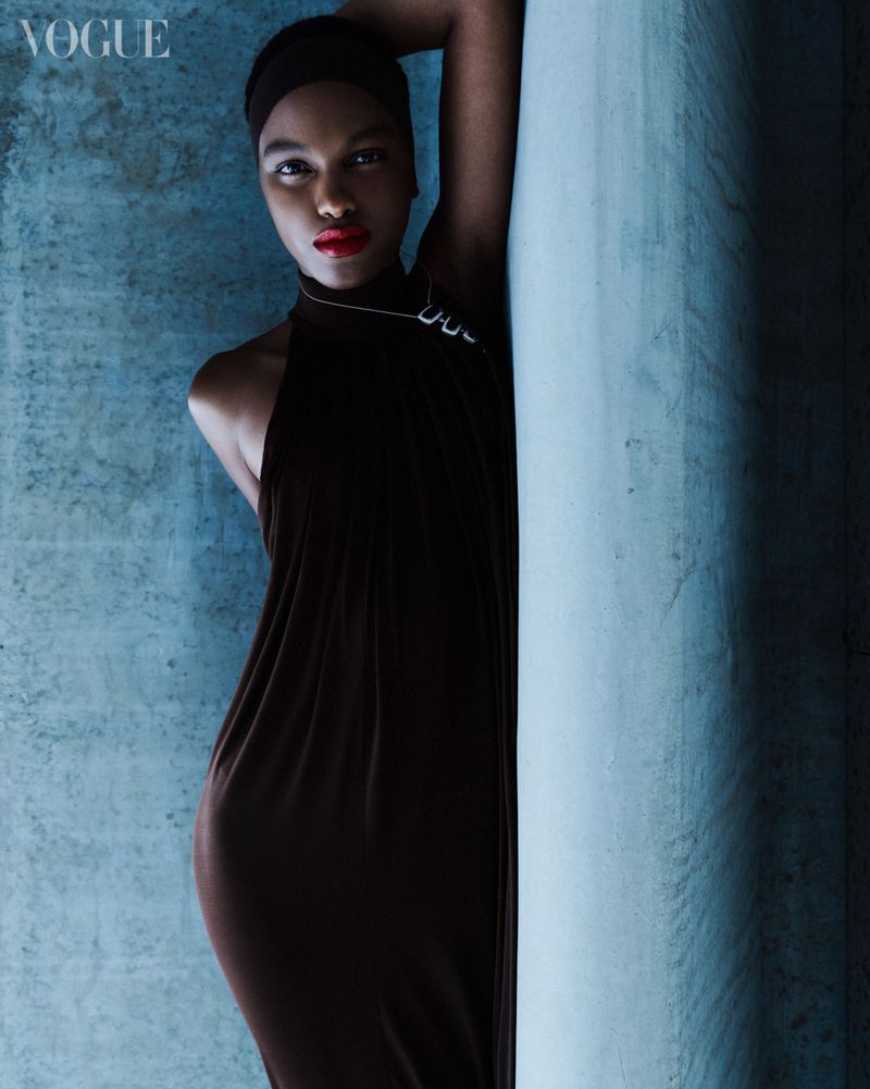 Elegant black woman in long black dress poses against textured blue wall in a fashion editorial. The model wears red lipstick.