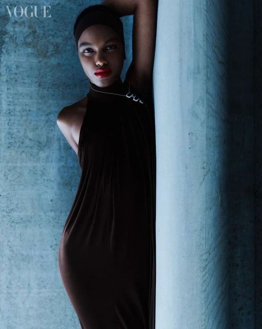 Elegant black woman in long black dress poses against textured blue wall in a fashion editorial. The model wears red lipstick.