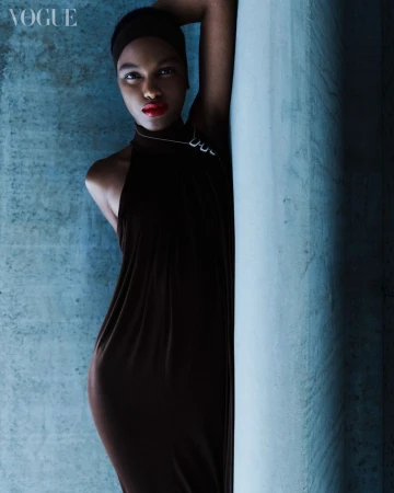 Elegant black woman in long black dress poses against textured blue wall in a fashion editorial. The model wears red lipstick.