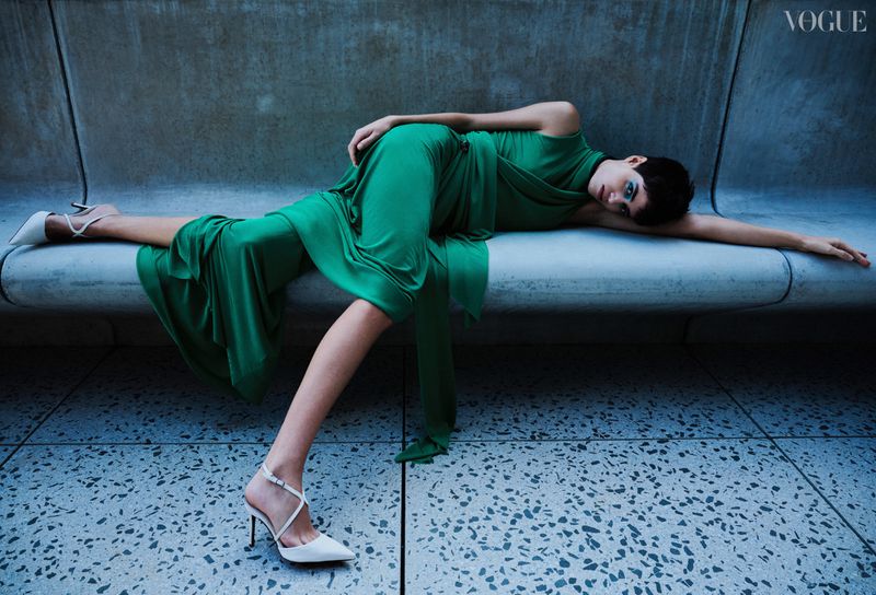 A fashion-style portrait of a woman in a green dress and high heels lying on a concrete structure, featuring a Vogue logo.