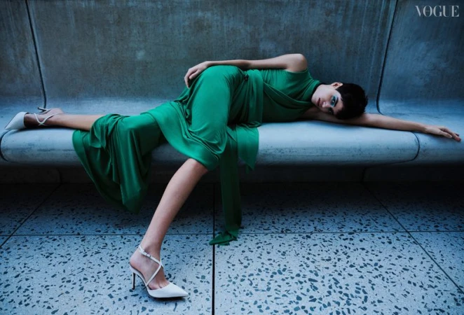 A fashion-style portrait of a woman in a green dress and high heels lying on a concrete structure, featuring a Vogue logo.