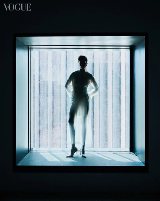 Adult female poses in sheer dress and stiletto heels, standing inside a brightly illuminated window frame for a fashion editorial.