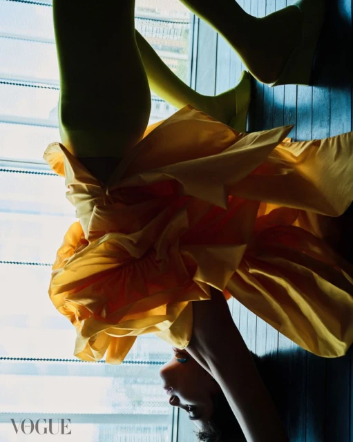 Fashionable woman poses in a yellow dress and lime-green tights near a window, showcasing a contemporary and stylish look, Vogue logo visible.