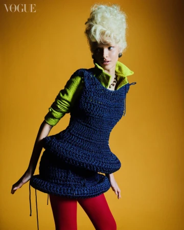 Fashion editorial shot of a female model with blonde hair wearing a dark blue knitted dress against a yellow backdrop in a studio.