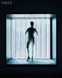 Model silhouetted standing in a large square window frame wearing a sheer white dress and heels against a backlit vertical slat pattern.