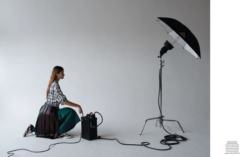 Woman in patterned top and pleated skirt kneels in a photo studio next to a black battery pack, with a studio light behind.