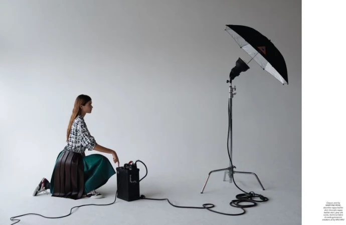 Woman in patterned top and pleated skirt kneels in a photo studio next to a black battery pack, with a studio light behind.