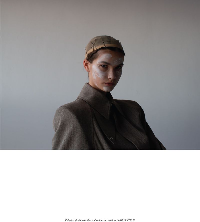 A portrait of a woman in a sharp-shouldered car coat by Phoebe Philo, wearing a head covering and face paint in a studio setting.