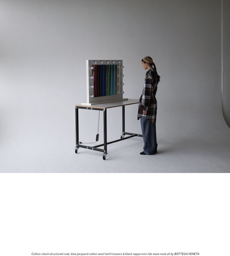 Woman in plaid coat and jeans stands near a table with colorful fabric display in a bright studio setting. Fashion editorial image.